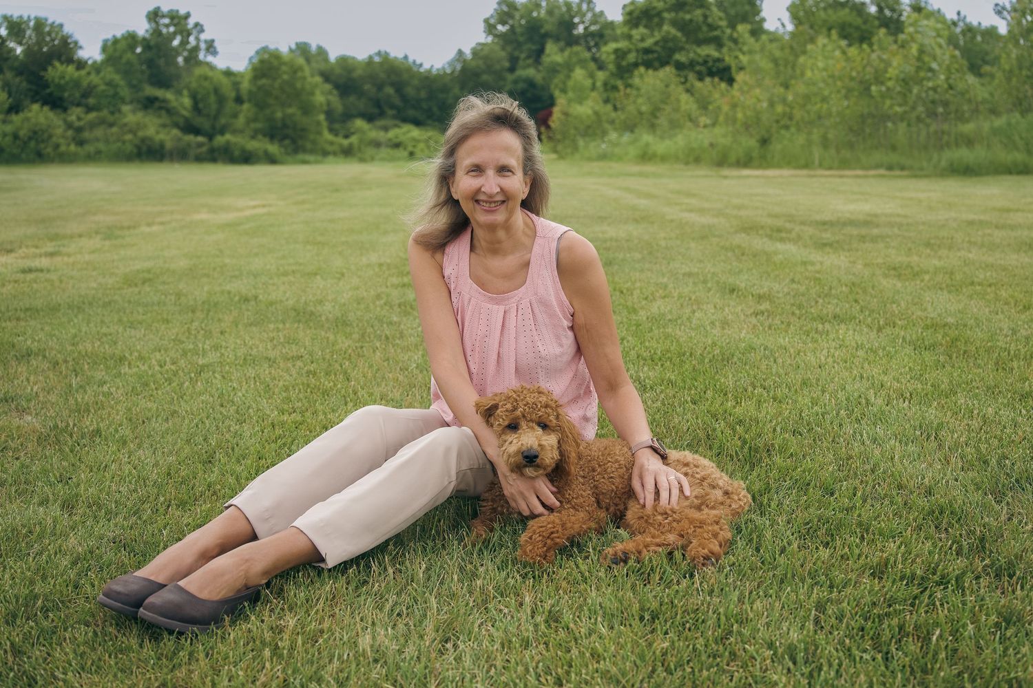 Woman in pink top sits on green grass with a fluffy red goldendoodle dog on a sunny day outdoors.