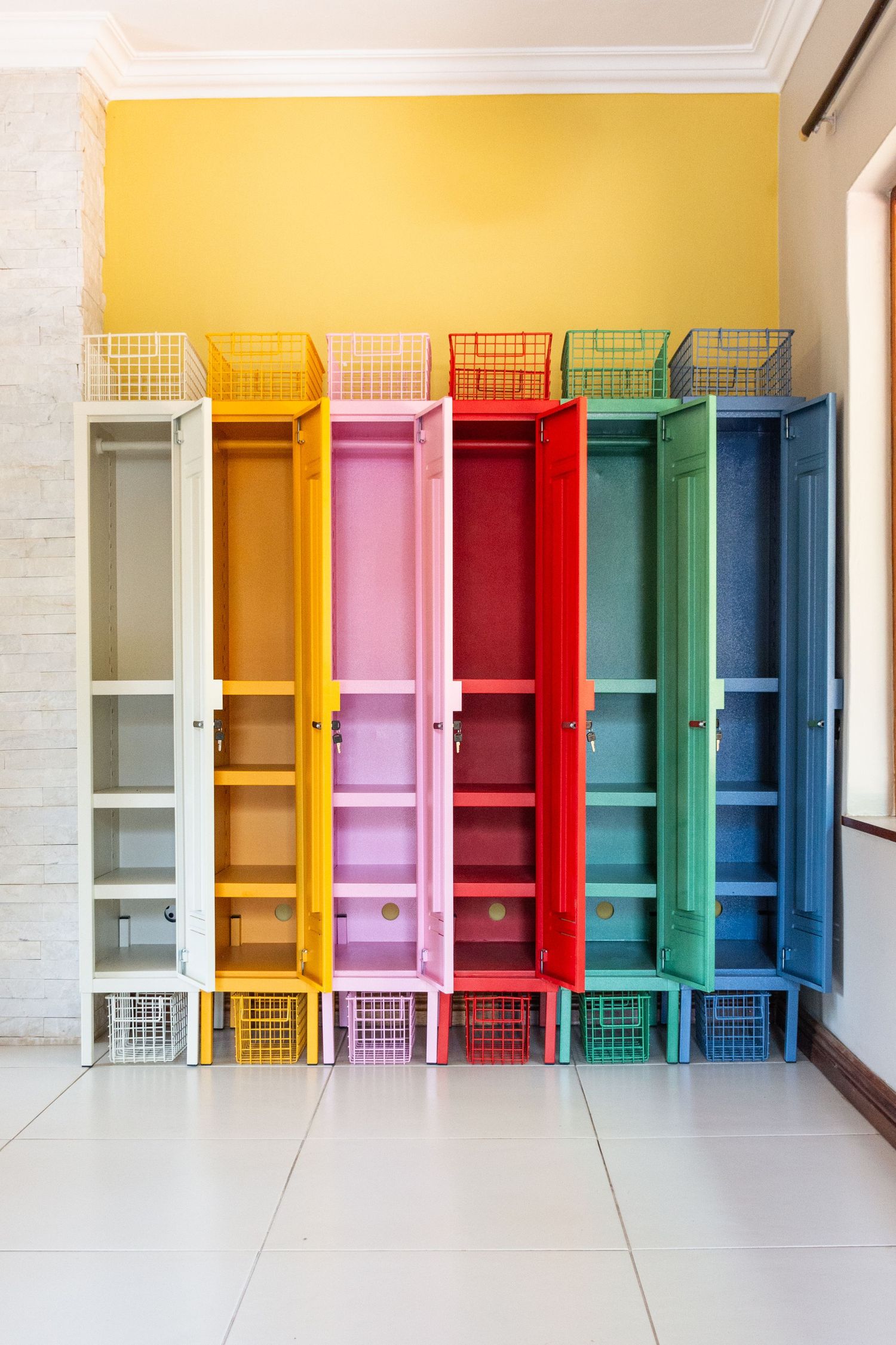 Colorful open lockers with wire baskets against a yellow wall, featuring rainbow hues including red, pink, blue, and green.