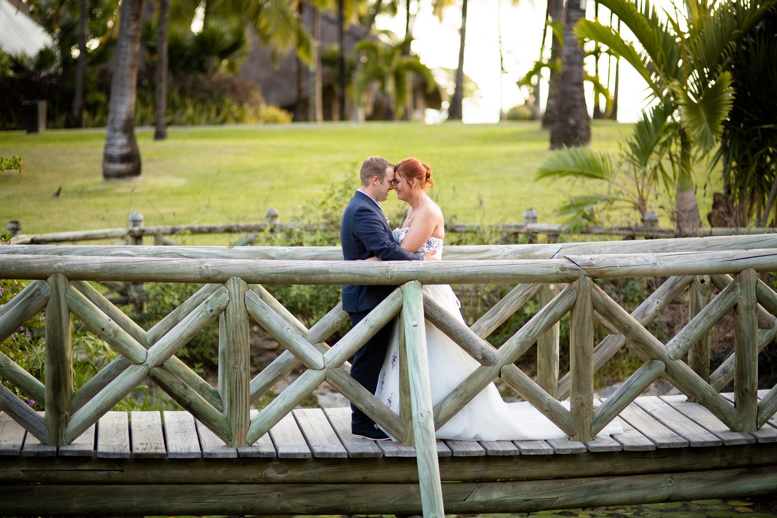 Bride and groom embracing on a wooden bridge surrounded by tropical palm trees and lush greenery.