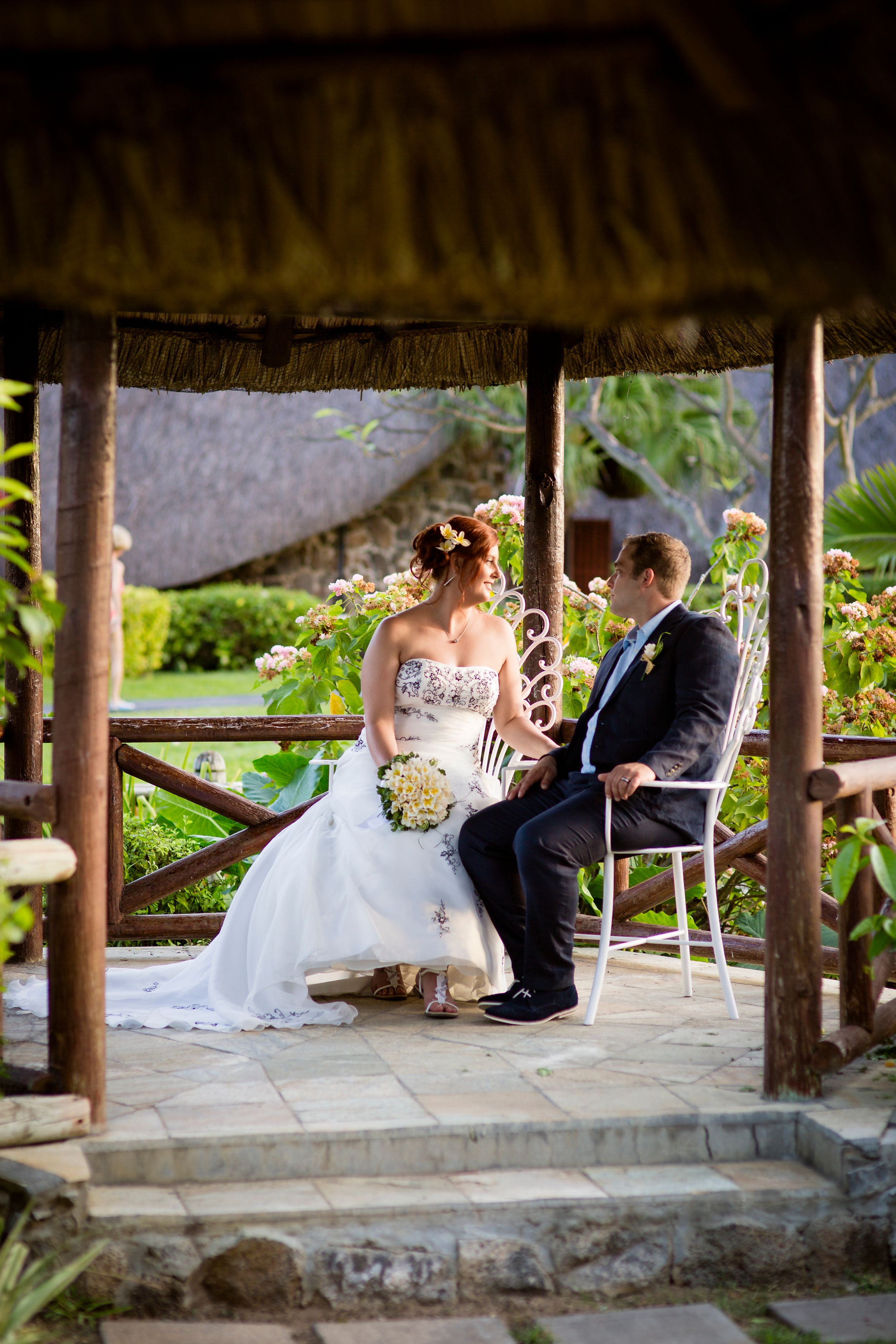 Bride and groom sitting together in an open-air thatched gazebo surrounded by tropical garden greenery.