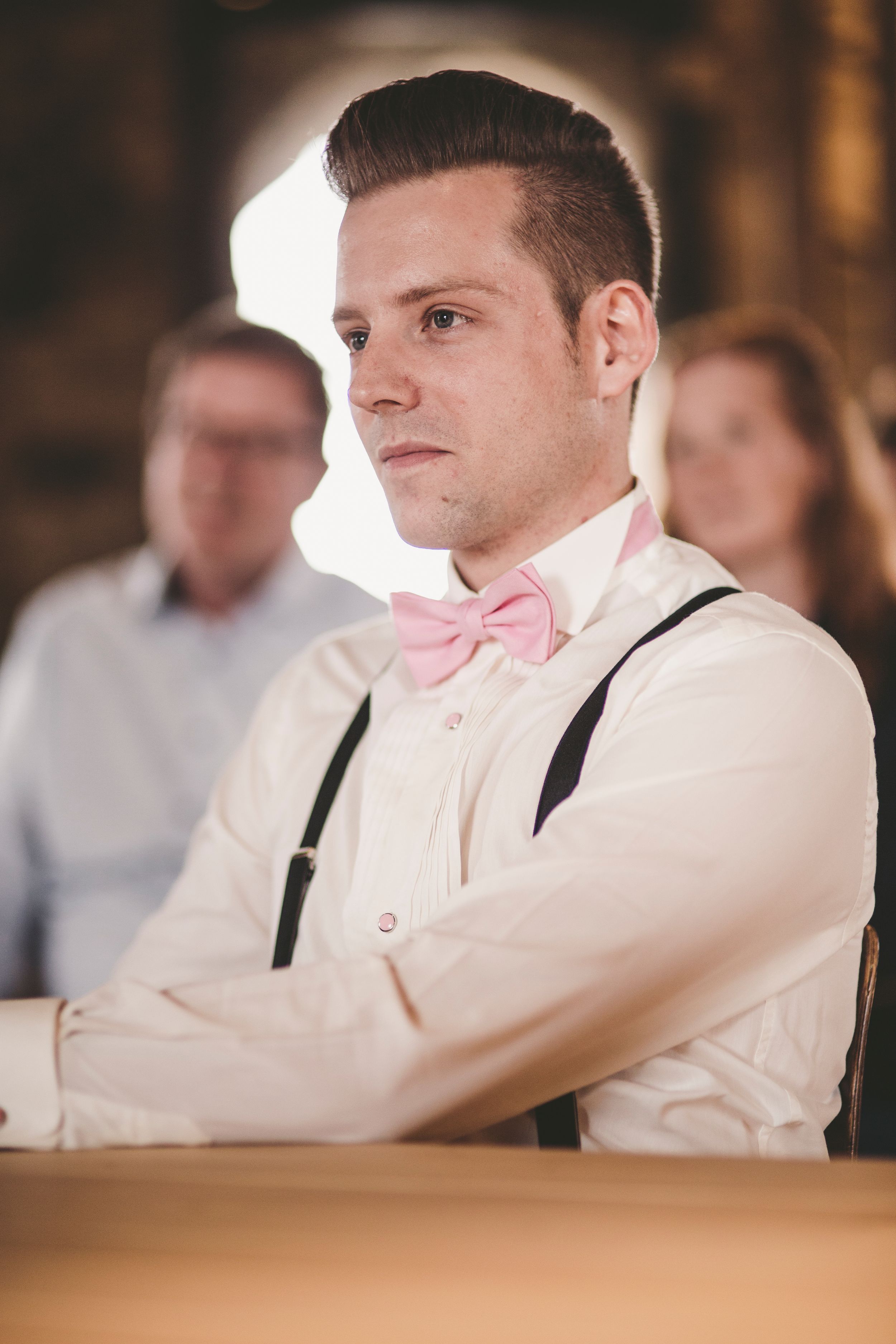 Groom wearing pink bow tie and suspenders sitting with arms crossed at a wedding ceremony.