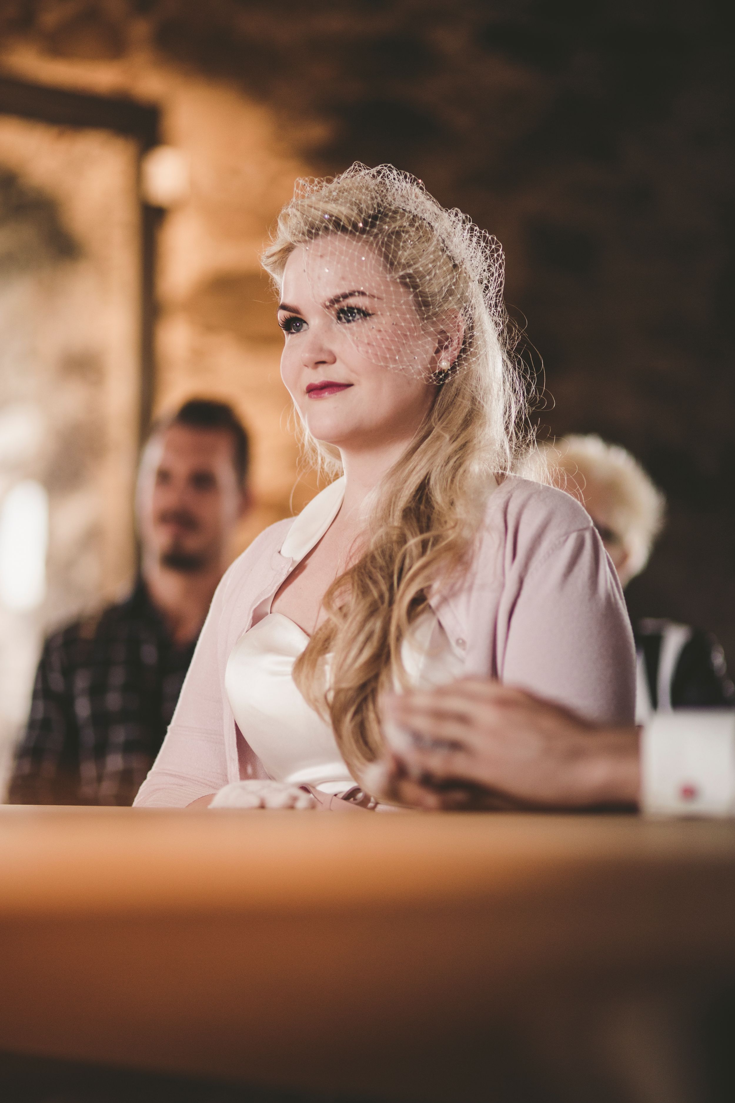Blonde bride wearing a birdcage veil and pink cardigan sitting at a rustic stone venue wedding.
