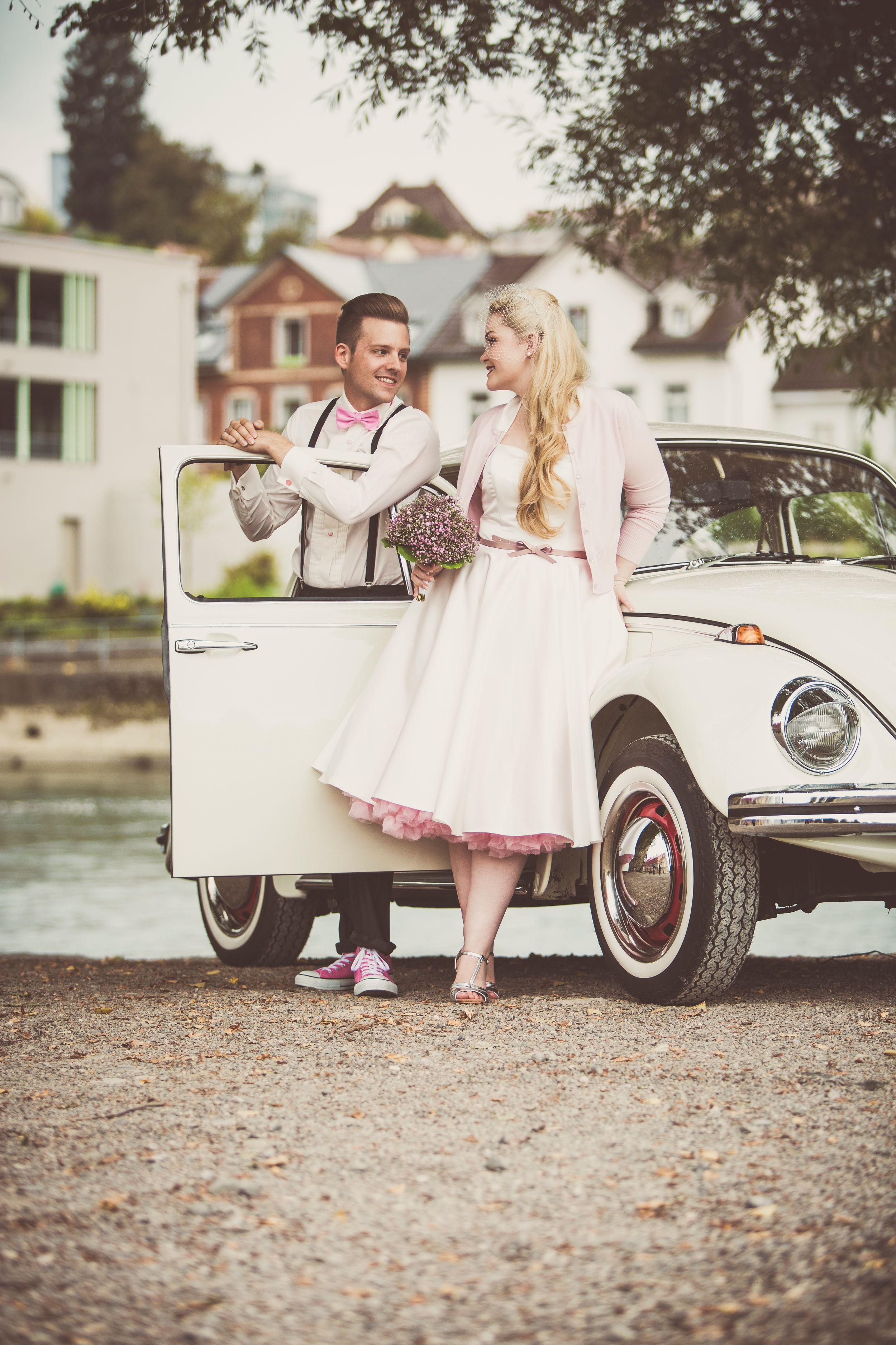 Bride in pink retro dress and groom with suspenders posing beside a vintage white VW Beetle car.