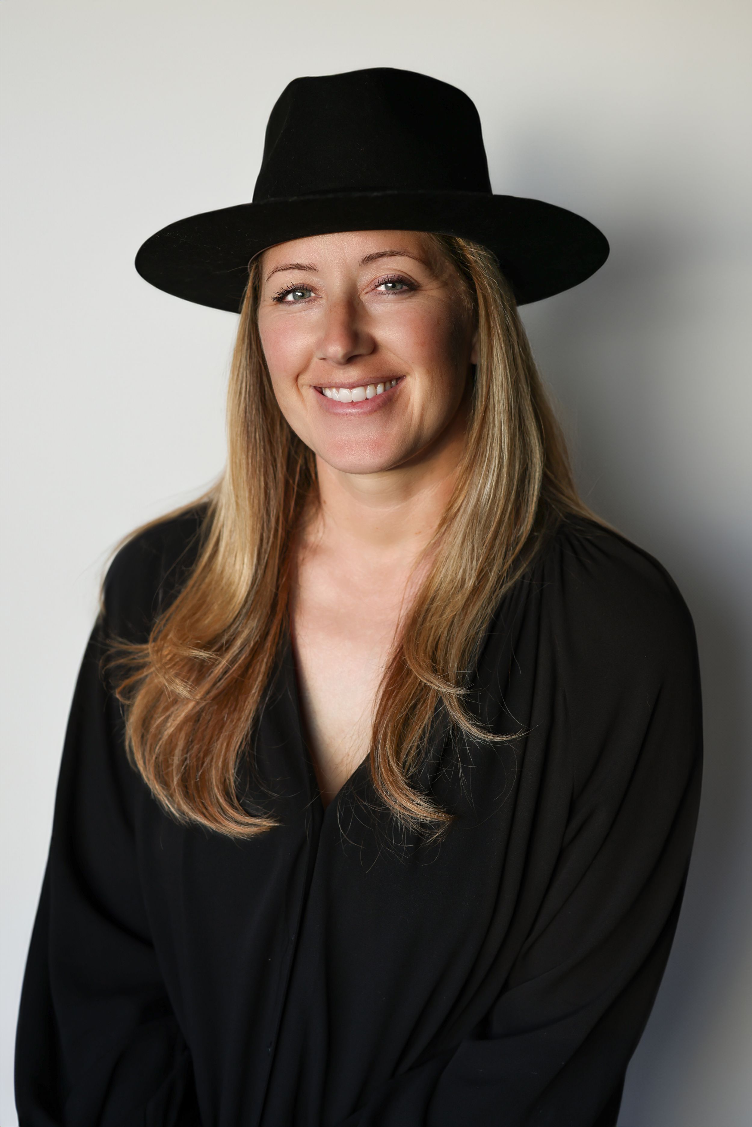 Smiling woman with long blonde hair wearing a black wide-brim hat and black top poses against a light gray background.