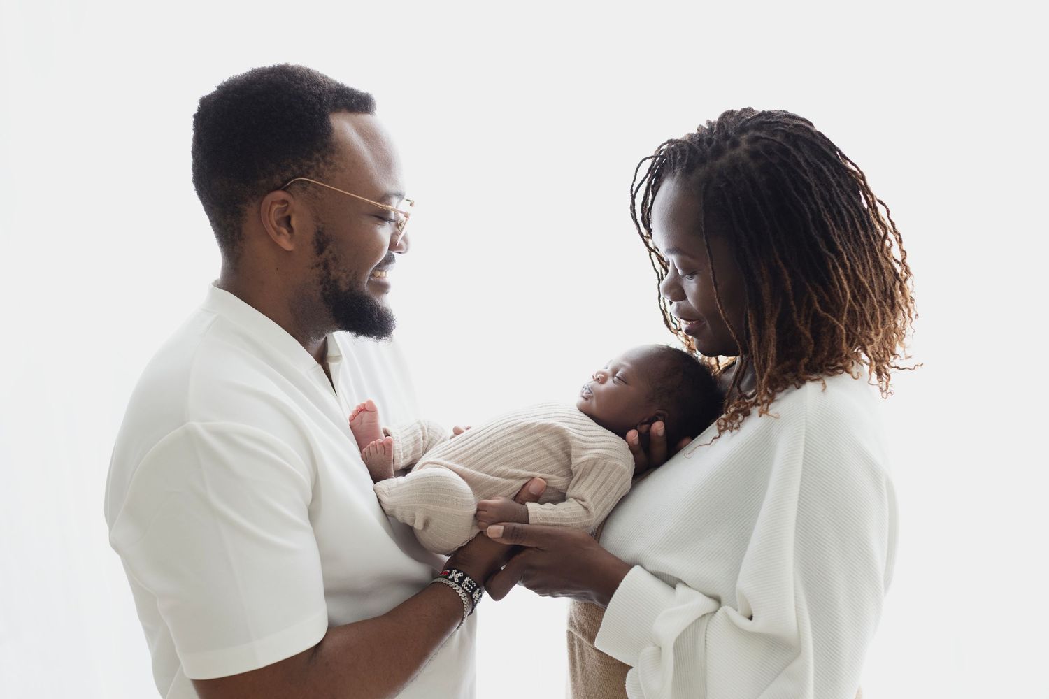 A Black couple in white outfits tenderly holds their newborn baby against a white studio background. Elizabeth Lang Photography Surrey