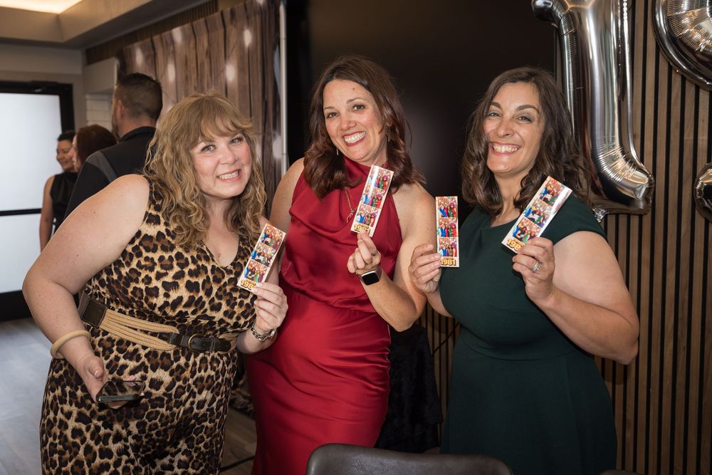 Three smiling women posing together at an event, holding photo booth strips as souvenirs.