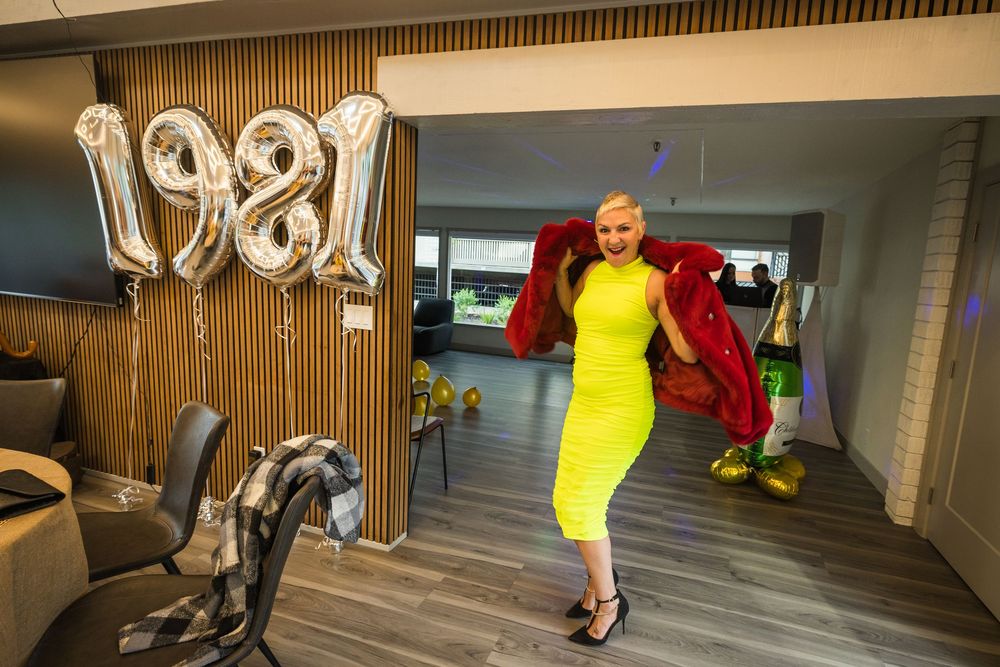 Woman in yellow dress holding red jacket poses by 1981 balloons in modern party venue.