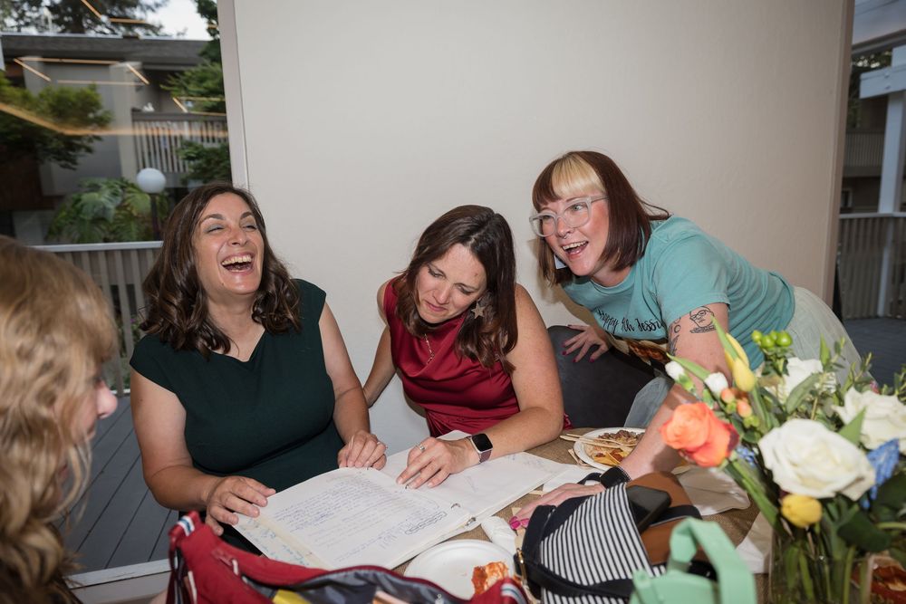 Three women laughing and sharing joyful moments together at a social gathering with flowers on the table.