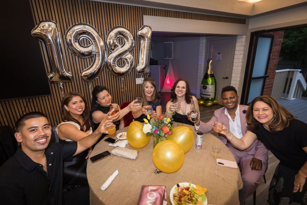 Group of friends celebrating a 1981 birthday party with champagne, gold balloons, and festive decorations at a restaurant.