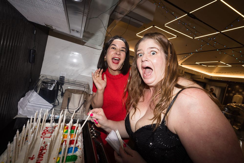 Two women posing excitedly near a birthday cake at a party, one in red dress, one in black top.