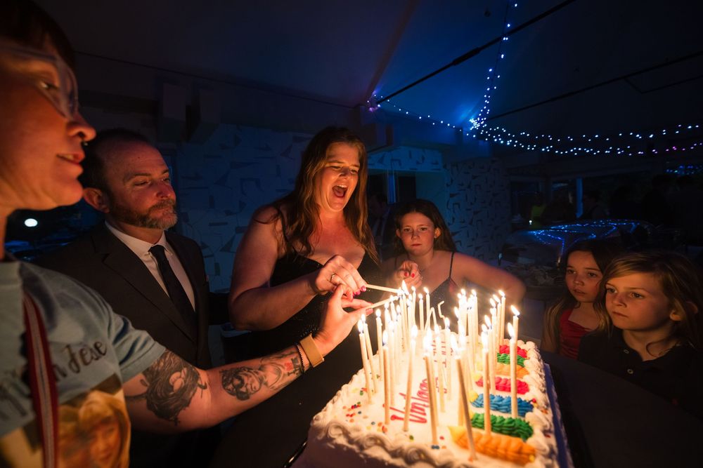 Group of people lighting candles on a large white birthday cake in a dimly lit party venue.
