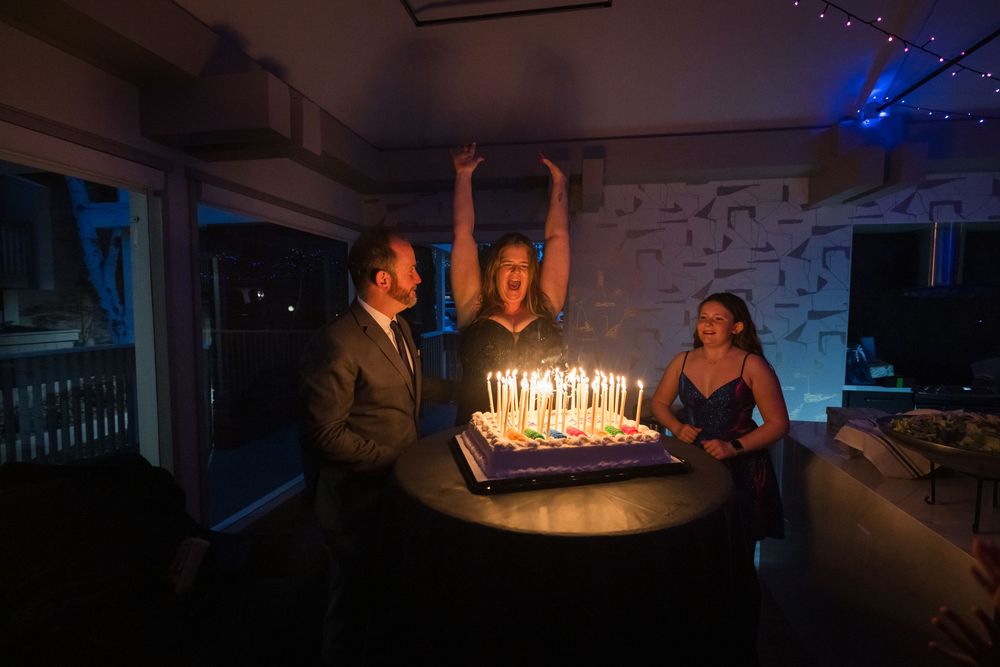 Three people celebrating a birthday with a candle-lit cake in a dimly lit room, one person raising their arms in excitement.