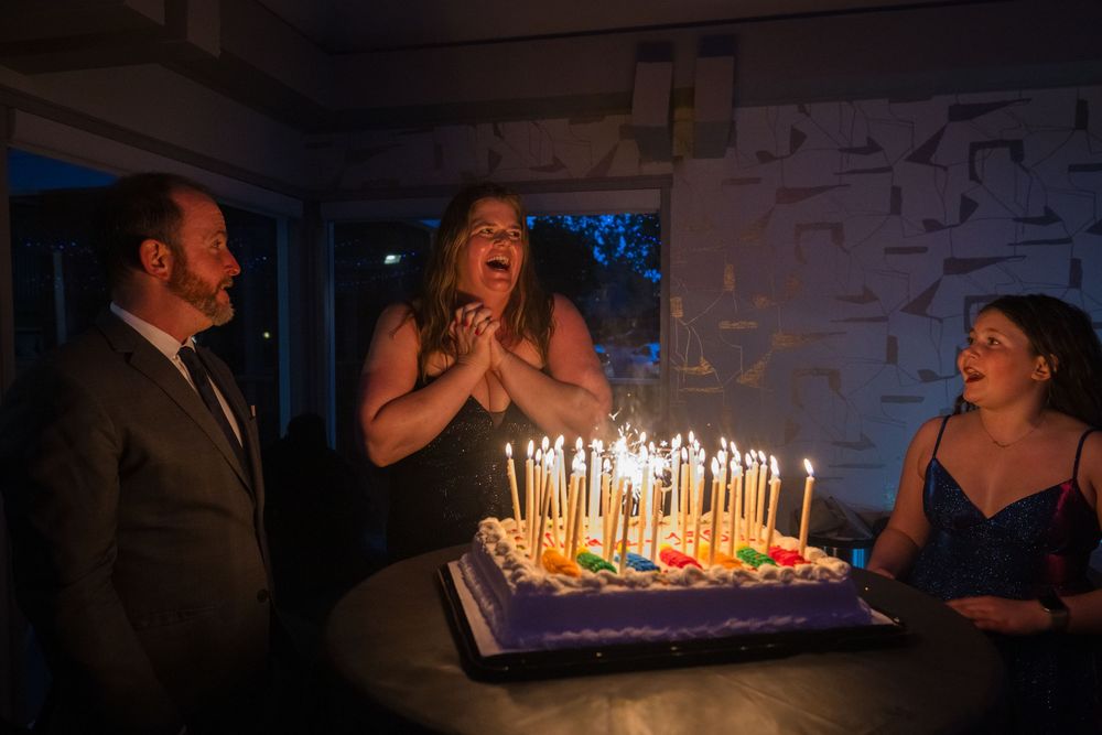 Group of people celebrating a birthday with a colorful cake lit with many candles in a dimly lit room.
