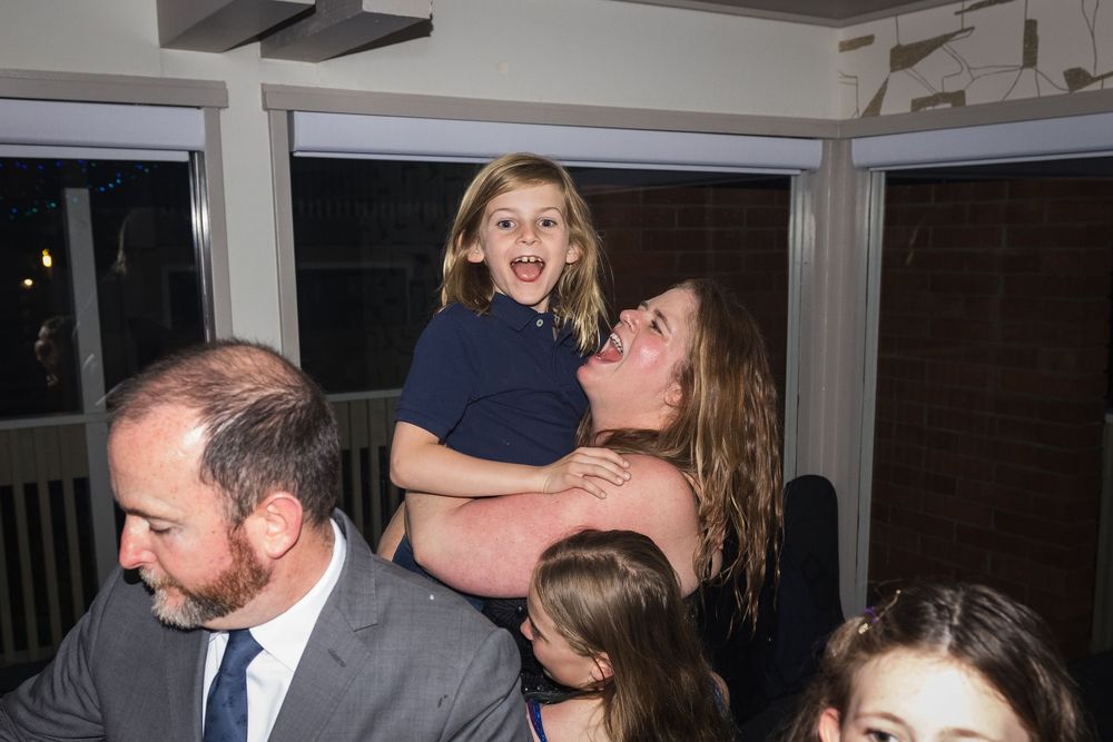 A woman joyfully lifts a laughing young girl at an indoor gathering, with other guests visible nearby.