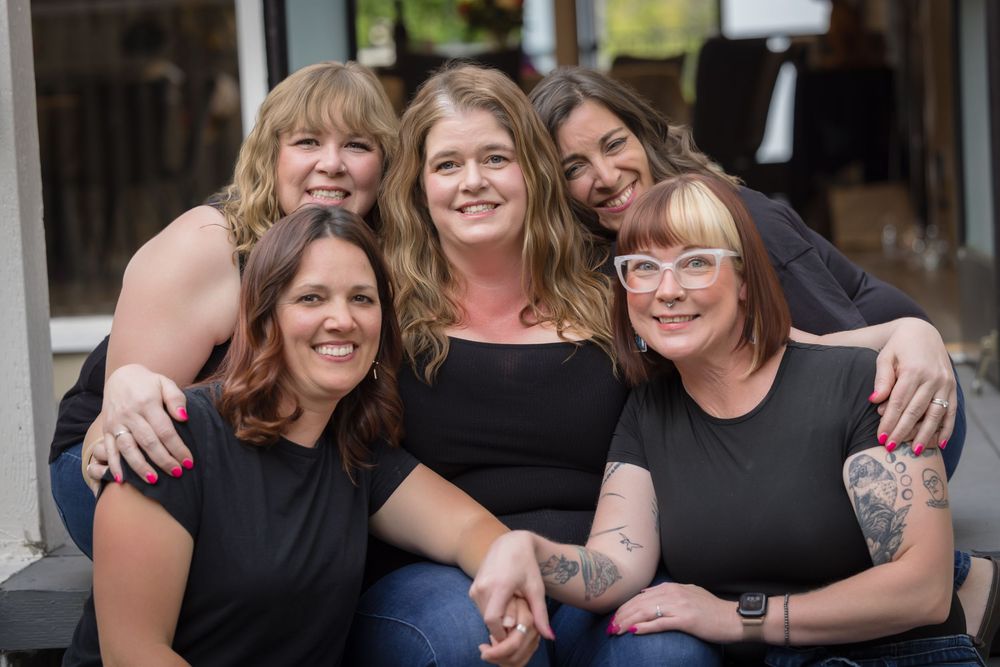 Five women smiling and posing together outdoors, wearing black tops, in a warm and friendly group portrait.