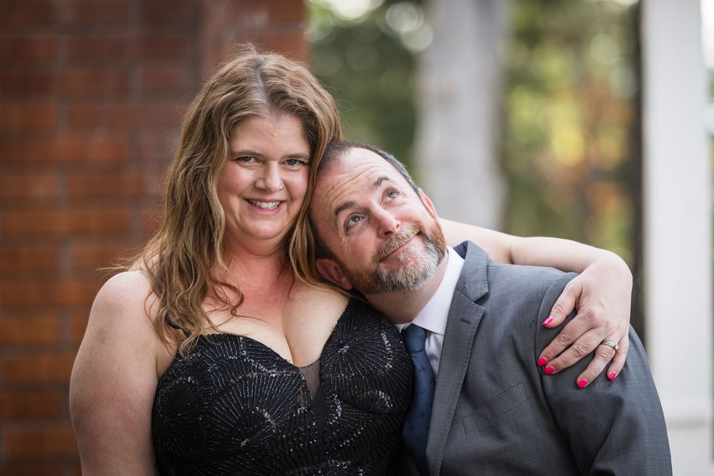 Smiling couple posing together, woman in black sequin dress and man in gray suit, embracing outdoors.