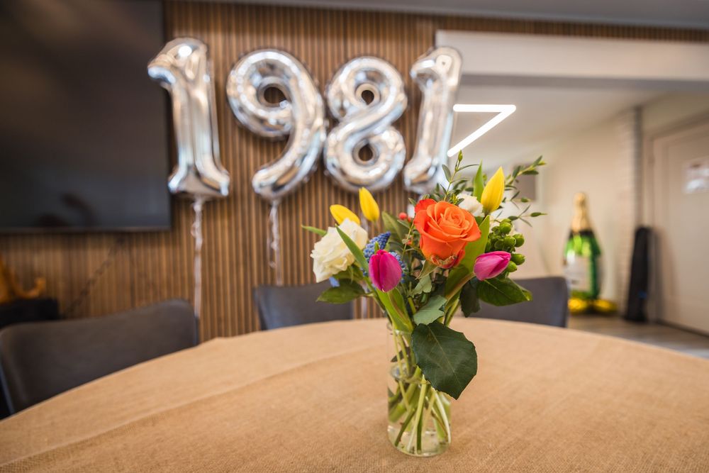 Colorful flower bouquet in glass vase on table with 1981 silver balloon numbers and champagne bottle in background.