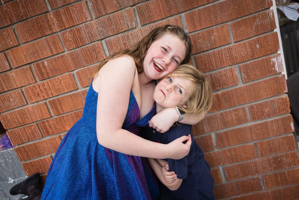 Two girls in blue dresses laughing and hugging against a red brick wall.