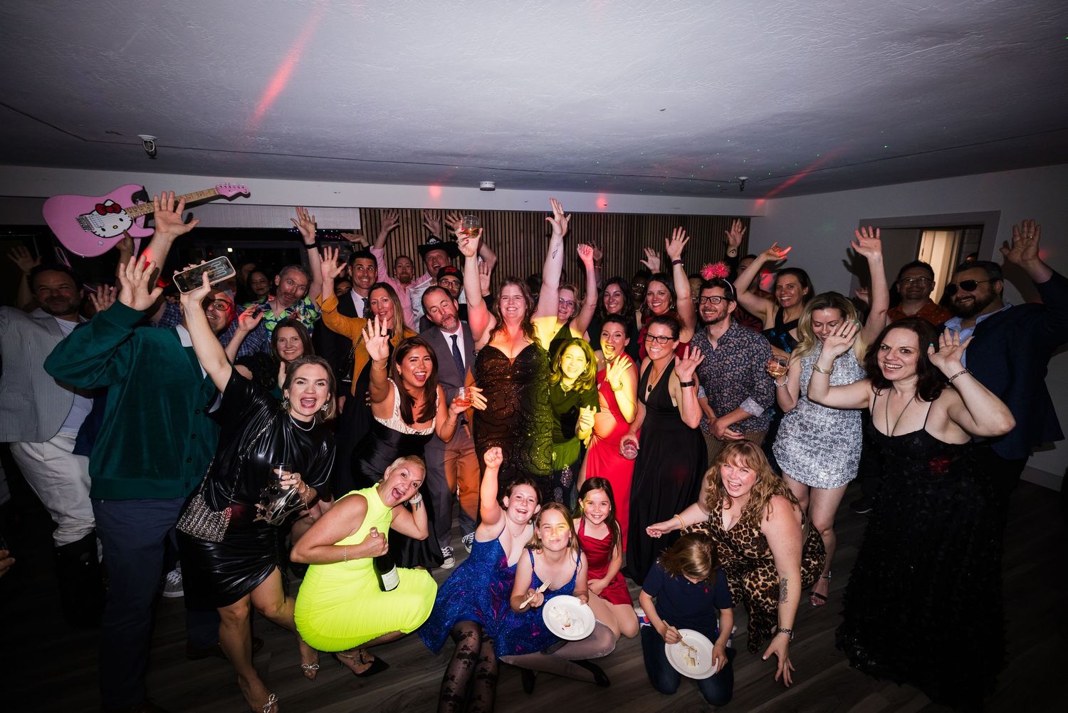 Large group of party guests celebrating together, raising hands and smiling at a festive indoor event.