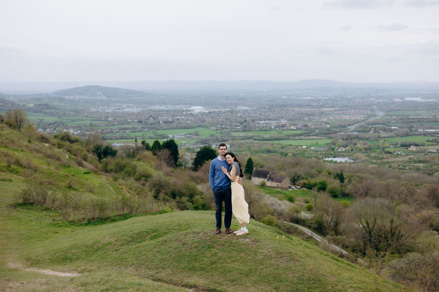 Couple embracing on a green hilltop with a panoramic view of a lush valley and distant mountains on a cloudy day.