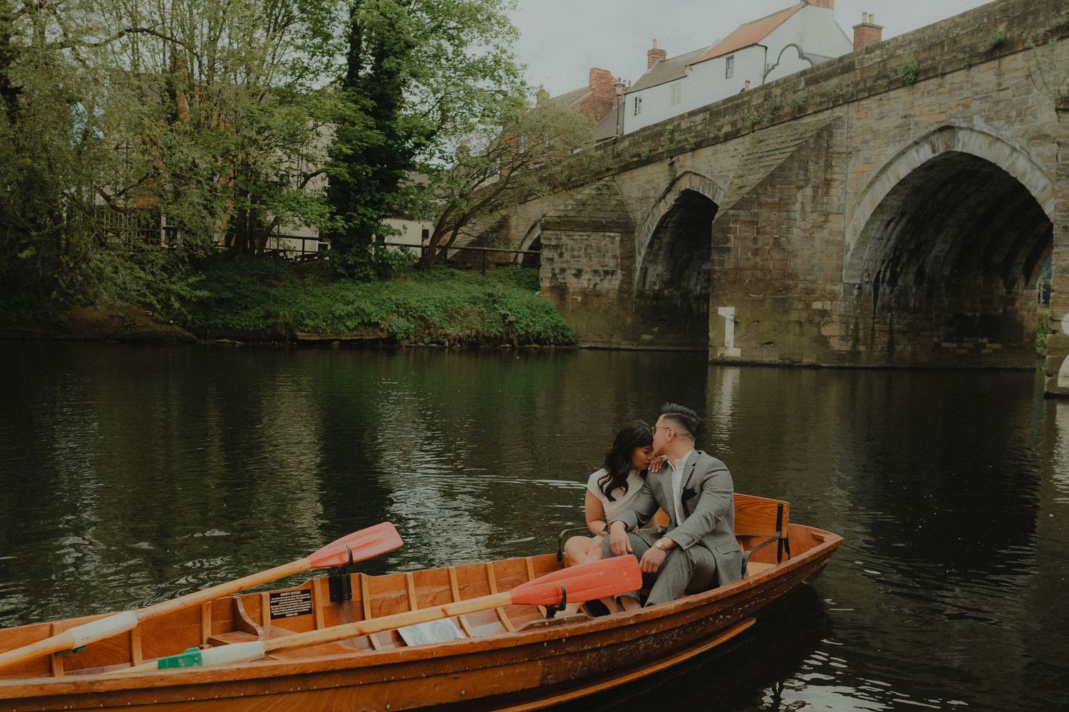 Couple sharing a romantic kiss on a charming cobblestone street beside historic stone buildings and lush green trees.