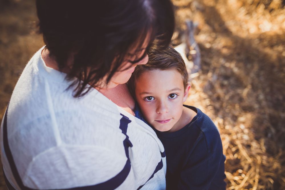 A young boy with blue eyes rests his head on a woman's shoulder outdoors in warm golden light.