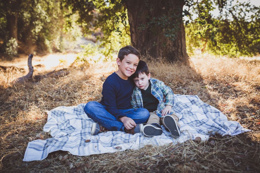 Two young boys sitting together on a blanket under a tree in a sunny outdoor setting.