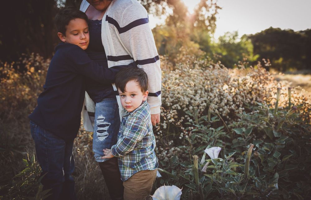 Two young boys hugging their father in a sunlit field with wildflowers during a family outdoor photo session.