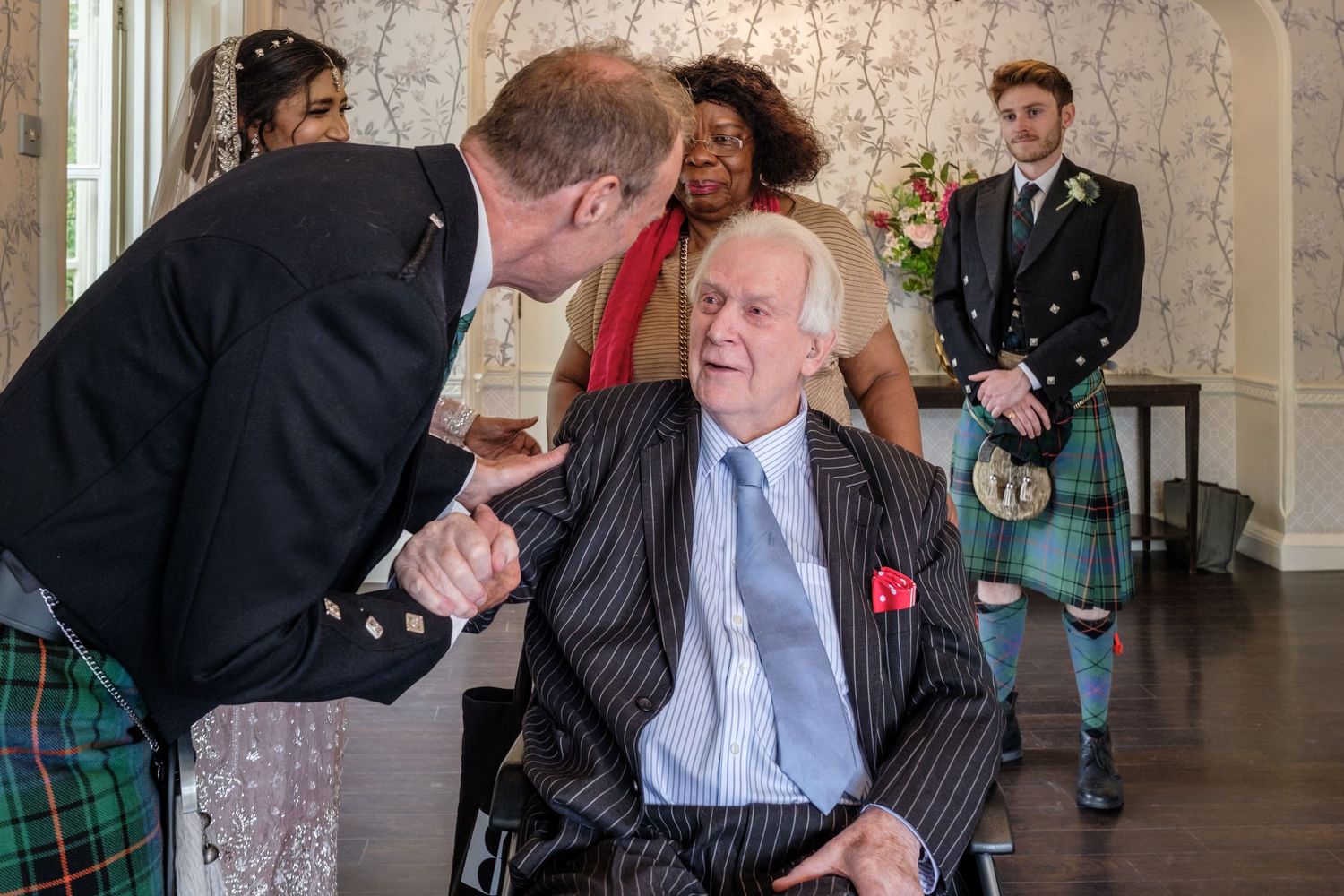 Bride in pink embroidered gown wipes tears while groom in Scottish kilt stands before her at elegant wedding ceremony.