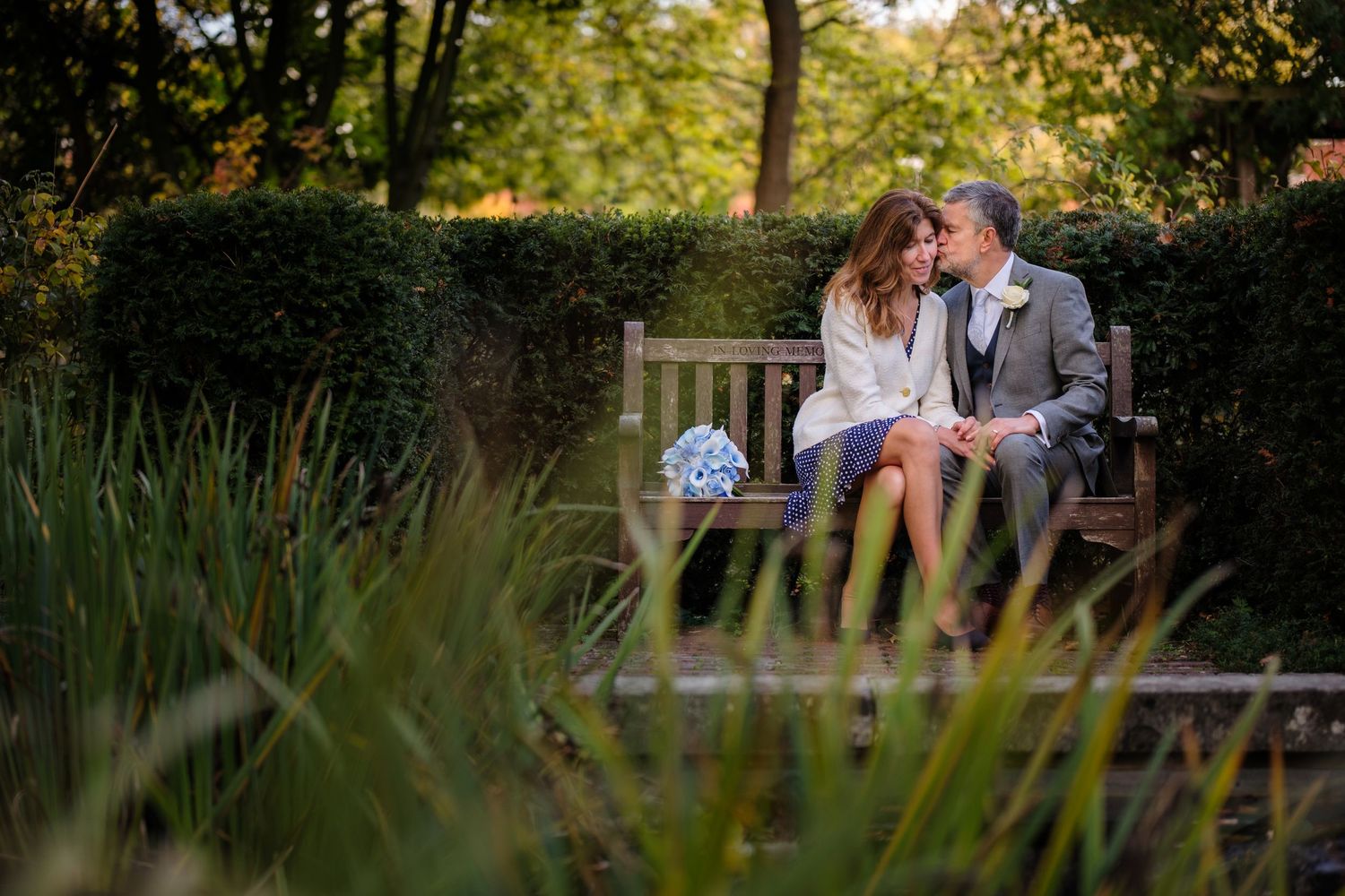 Couple sitting closely on a wooden bench in a lush garden, with a blue bouquet nearby.