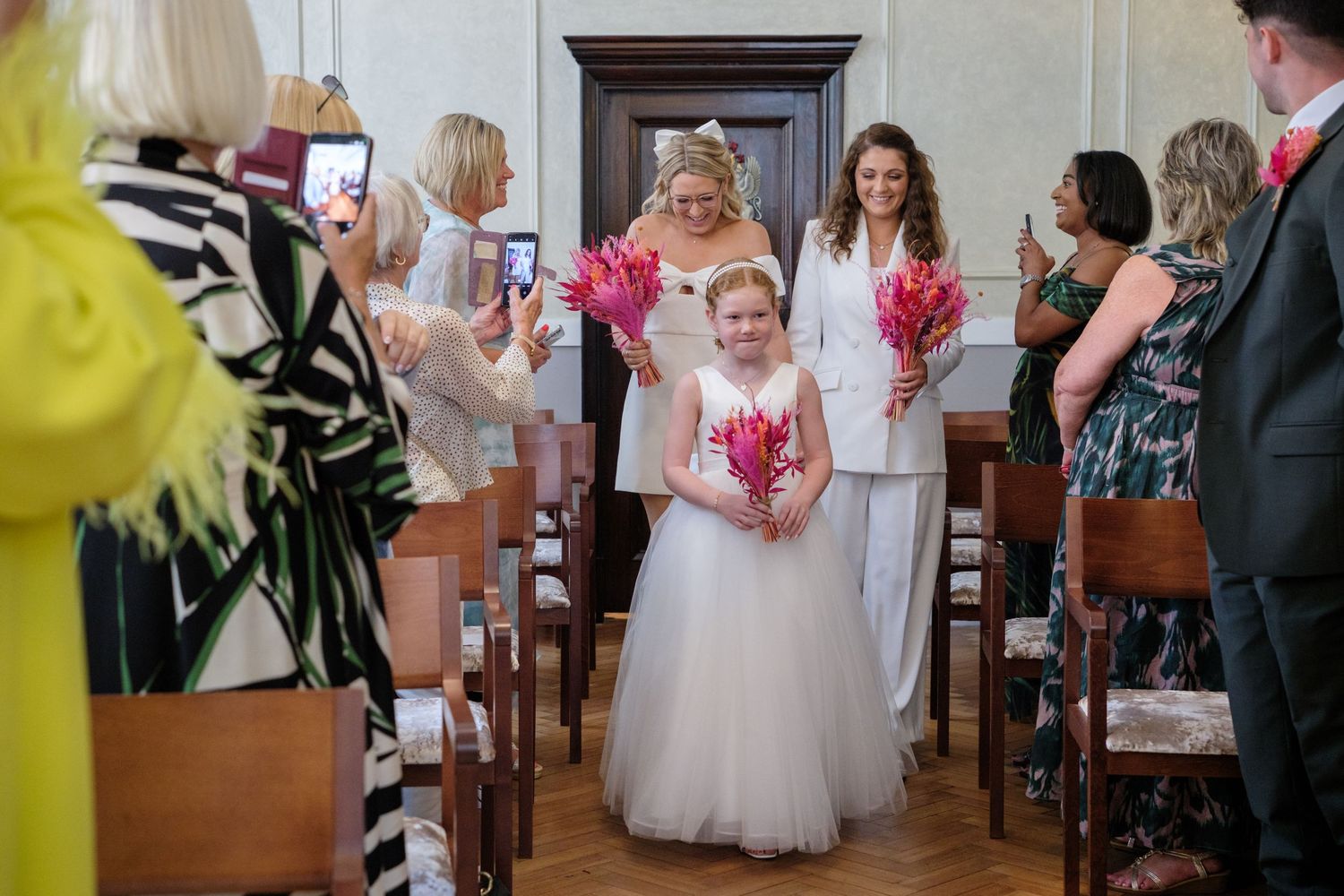 Bride walks down the aisle with flower girl holding pink bouquets at an elegant wedding ceremony.