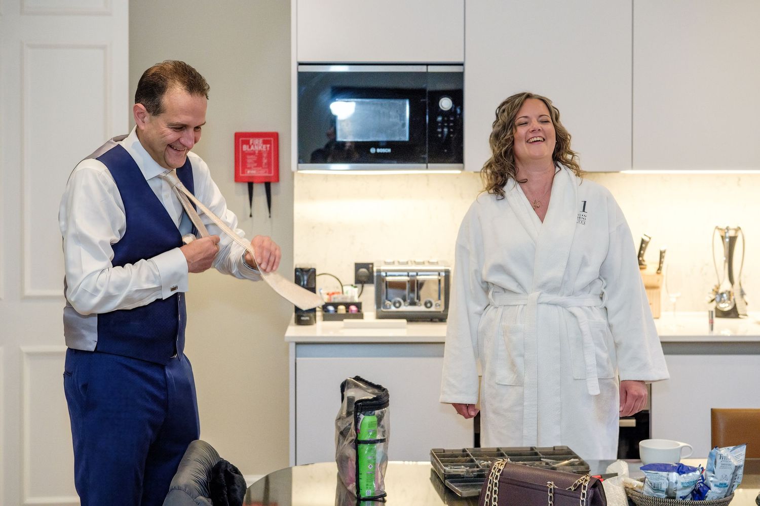 Man in blue vest and woman in white robe stand in modern hotel kitchenette, smiling and unpacking items.