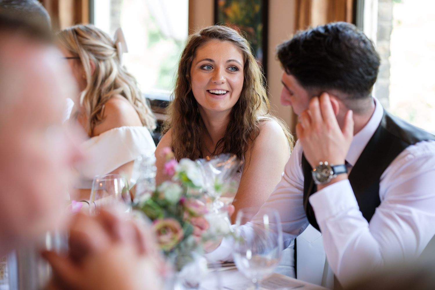 Bride smiling joyfully at wedding reception dinner table with guests and floral bouquet.
