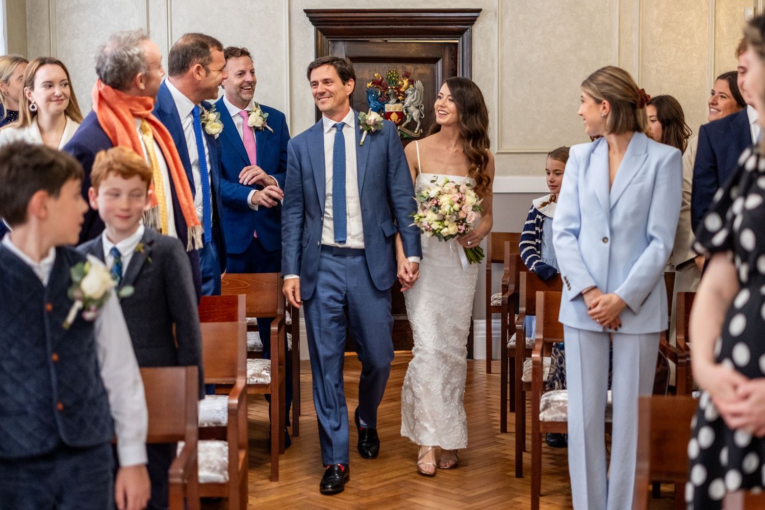 Bride and groom walking down the aisle at a bright indoor wedding ceremony with smiling guests.