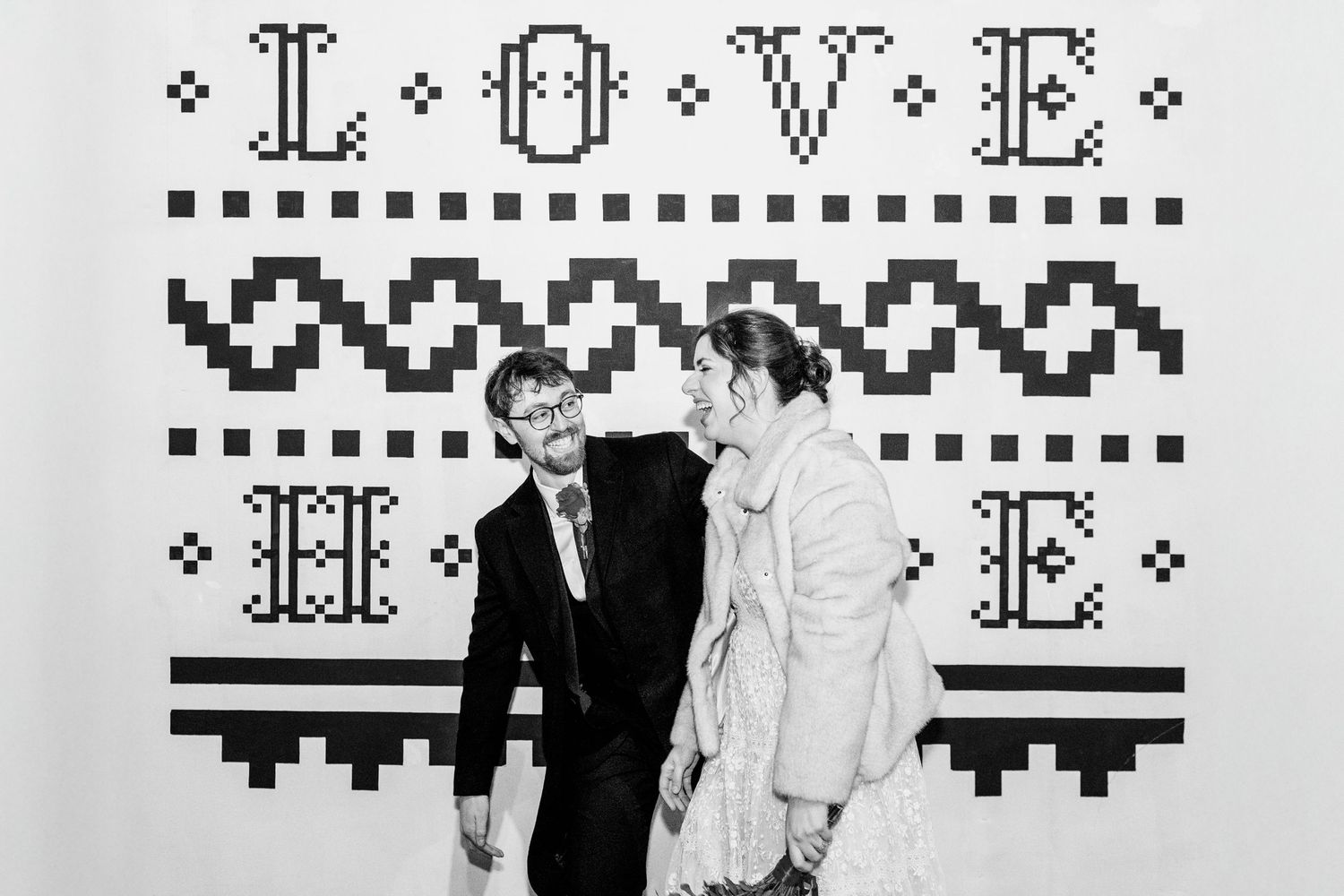 Black and white photo of a couple laughing in front of a decorative LOVE HOME cross-stitch wall backdrop.