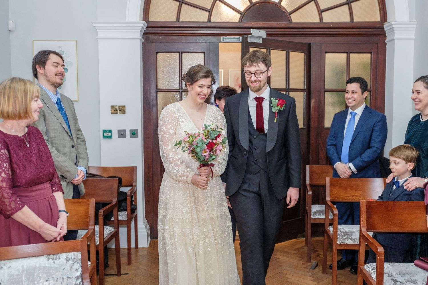 Bride and groom walk down the aisle smiling at a civil wedding ceremony with guests standing nearby.
