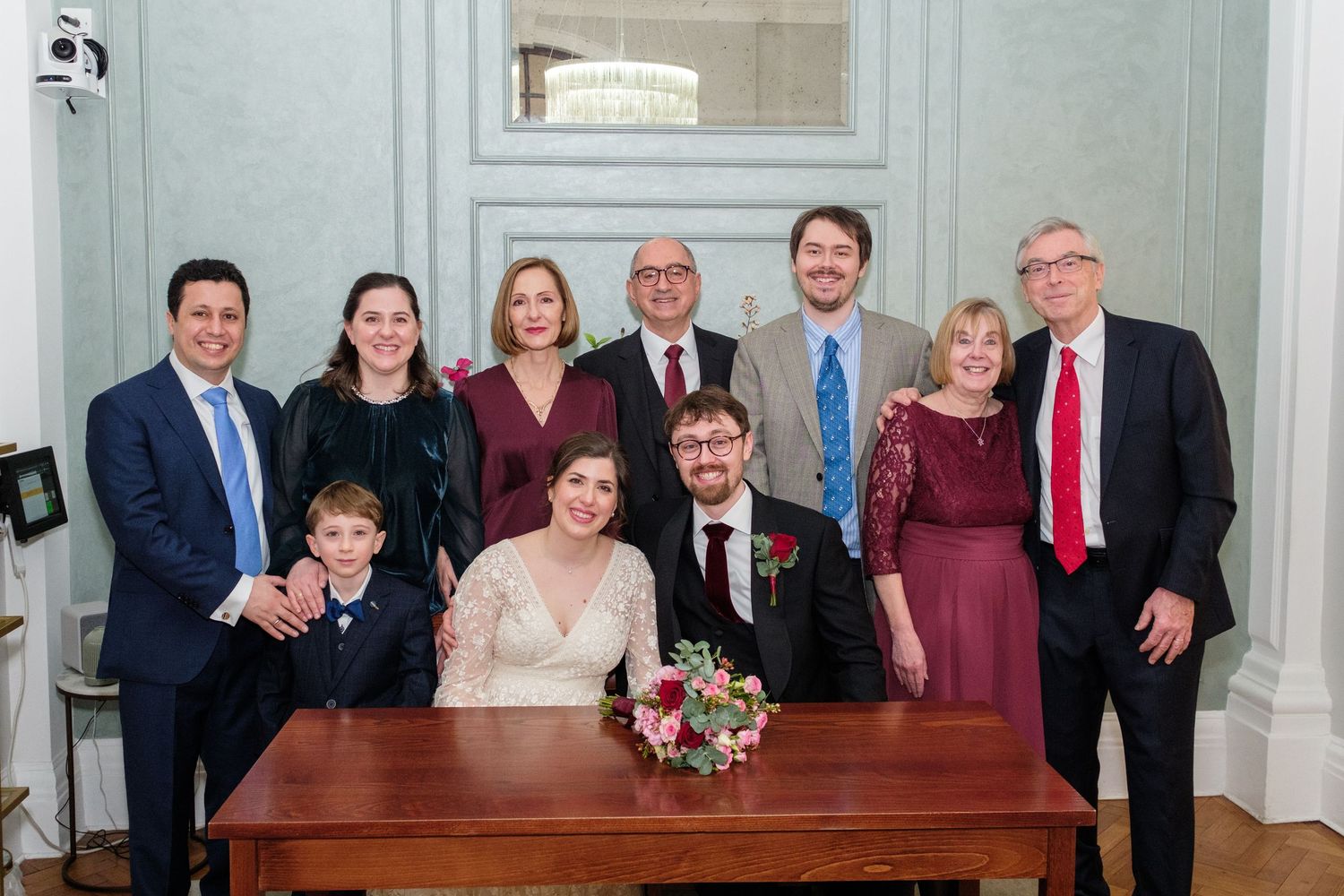 Family group photo at a wedding ceremony with bride and groom seated at signing table, surrounded by smiling relatives in formal attire.
