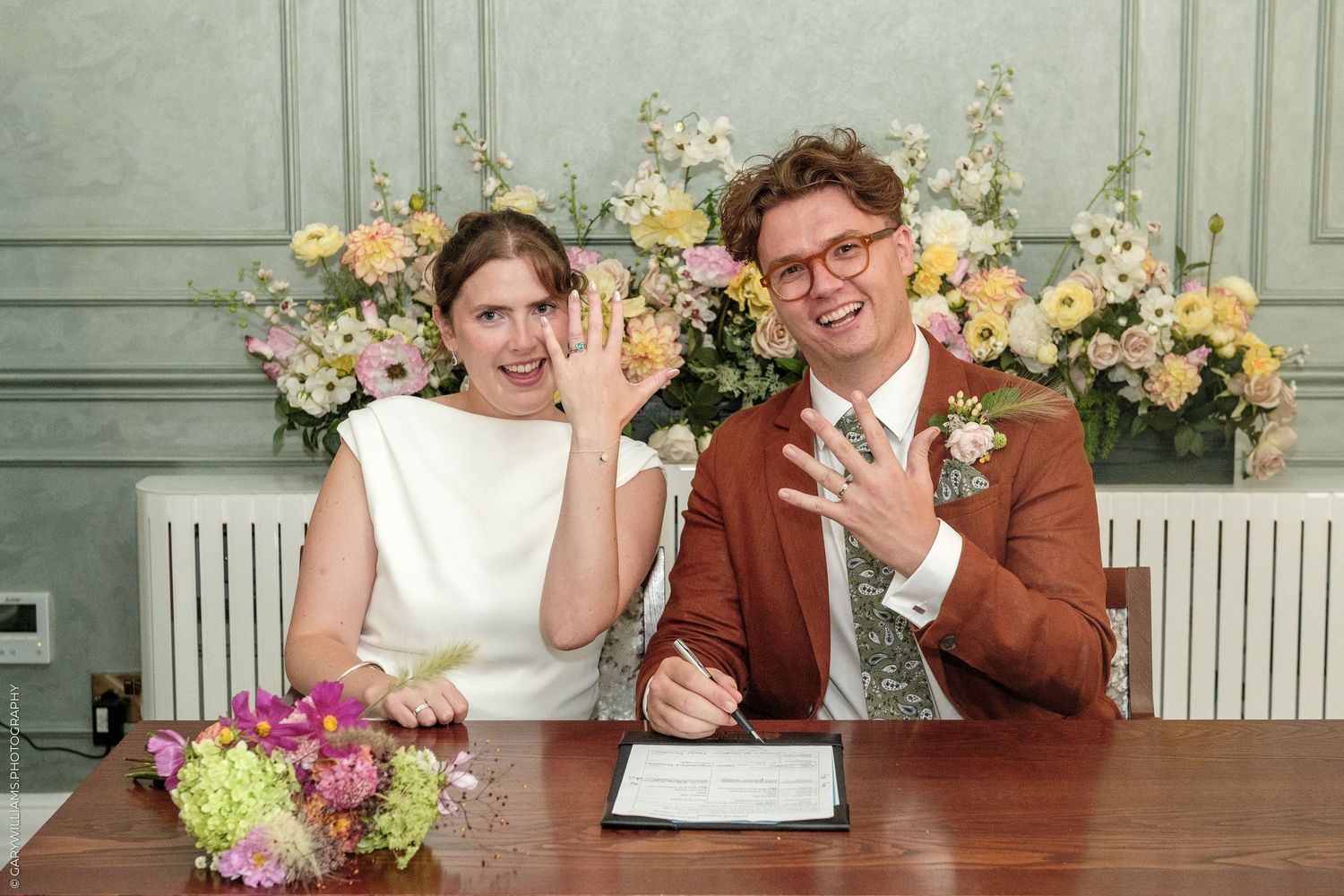 Couple signing wedding certificate at table, showing rings, surrounded by pastel floral arrangements in elegant venue.
