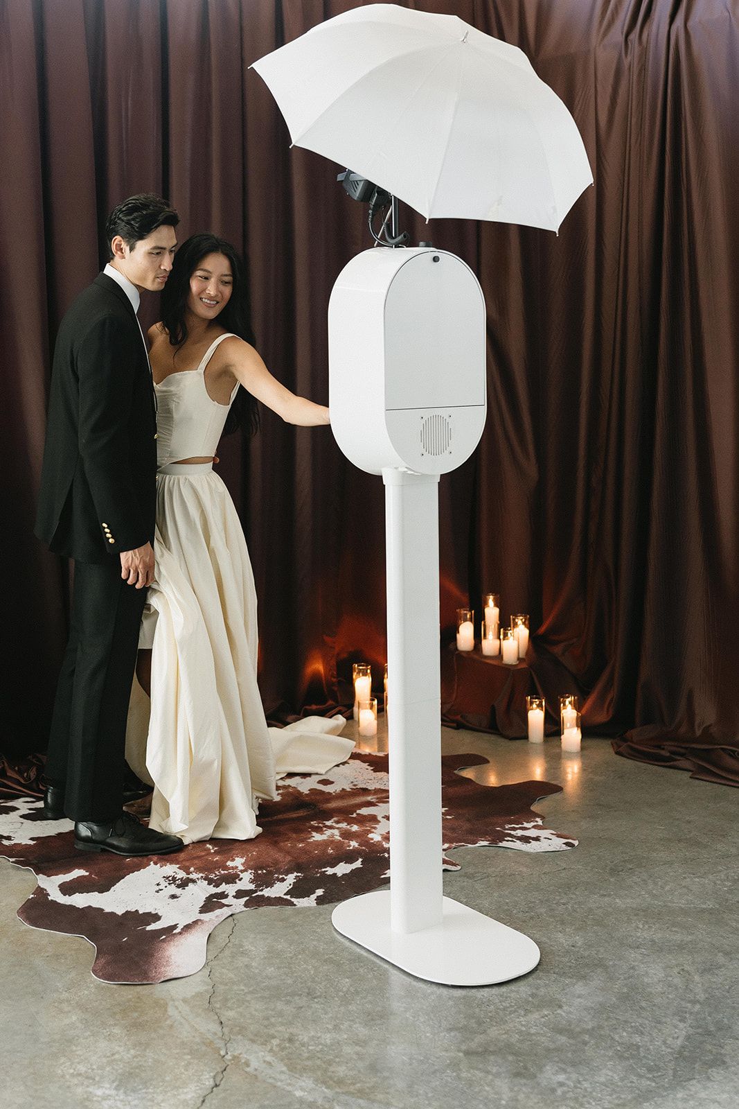 A couple in wedding attire poses beside a white photo booth kiosk with umbrella flash, set against brown drape backdrop.