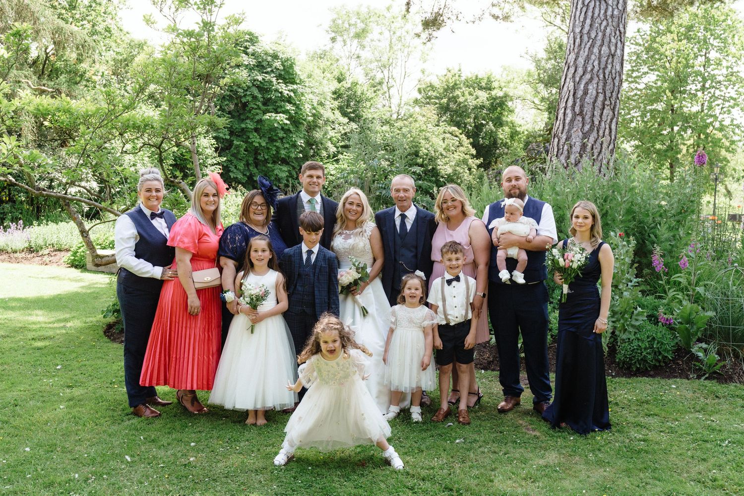 Wedding party group photo in a lush garden, bride and groom surrounded by family and flower girls.