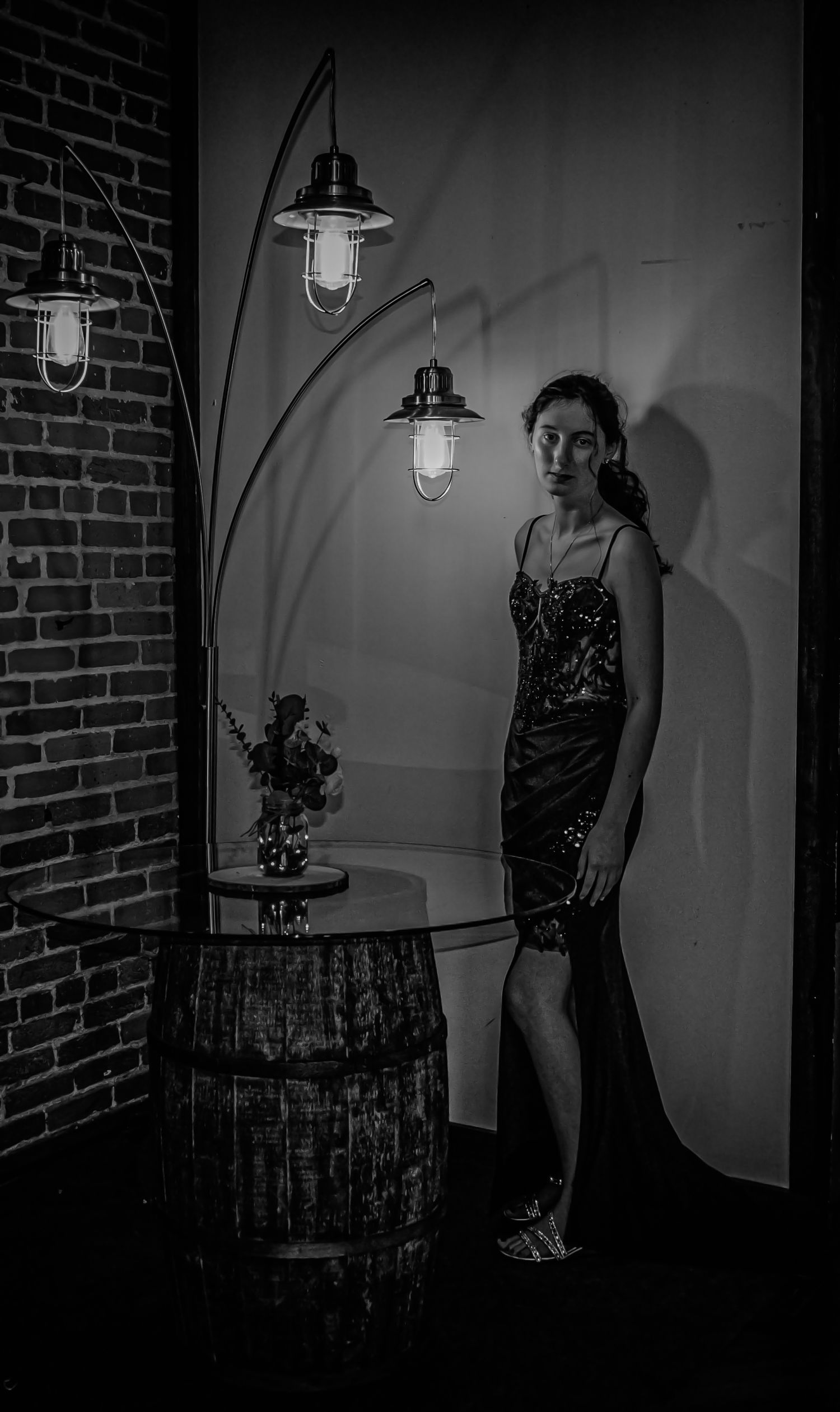 Woman in elegant gown poses beside barrel table and arc lamp in moody black and white interior setting.