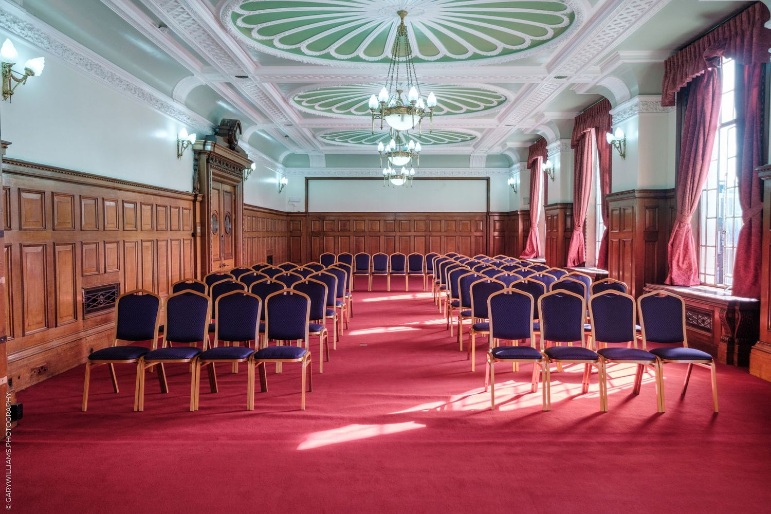 Elegant conference room with red carpet, blue chairs in rows, wood paneling, ornate ceiling, and chandelier lighting.
