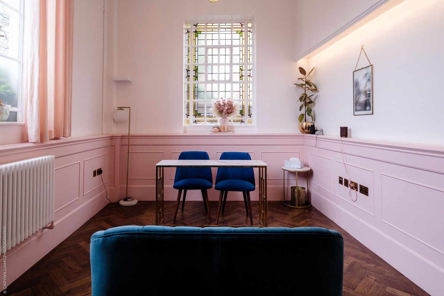 Pink dining room with blue velvet chairs, white table, herringbone wood floor, and teal sofa in foreground.
