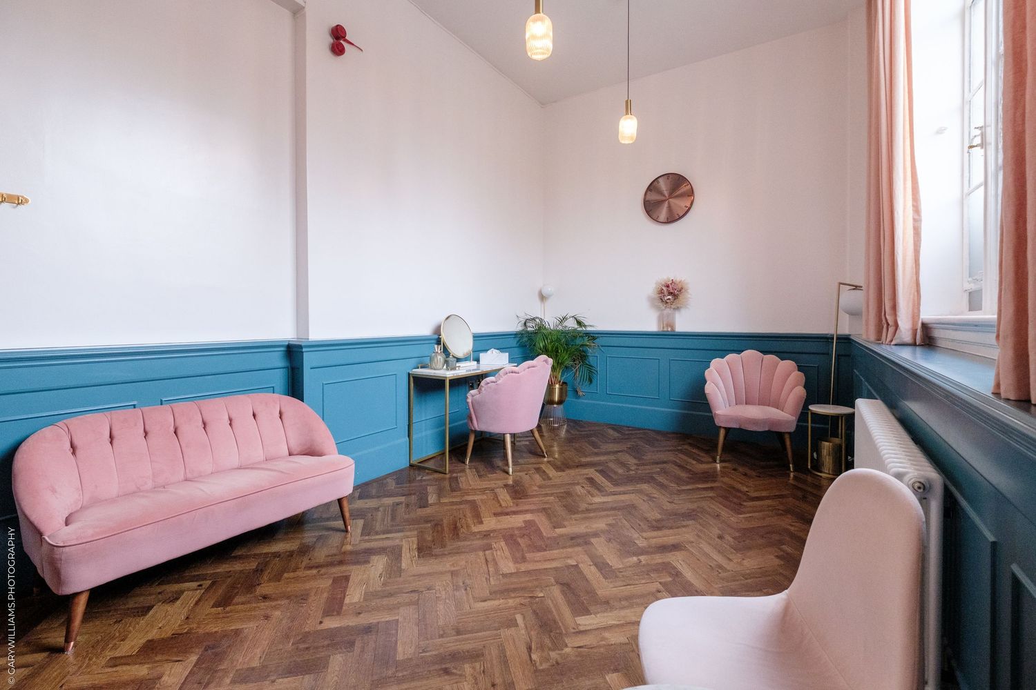Elegant waiting room with pink velvet sofa and chairs, teal wainscoting, herringbone parquet floor, and gold vanity desk.