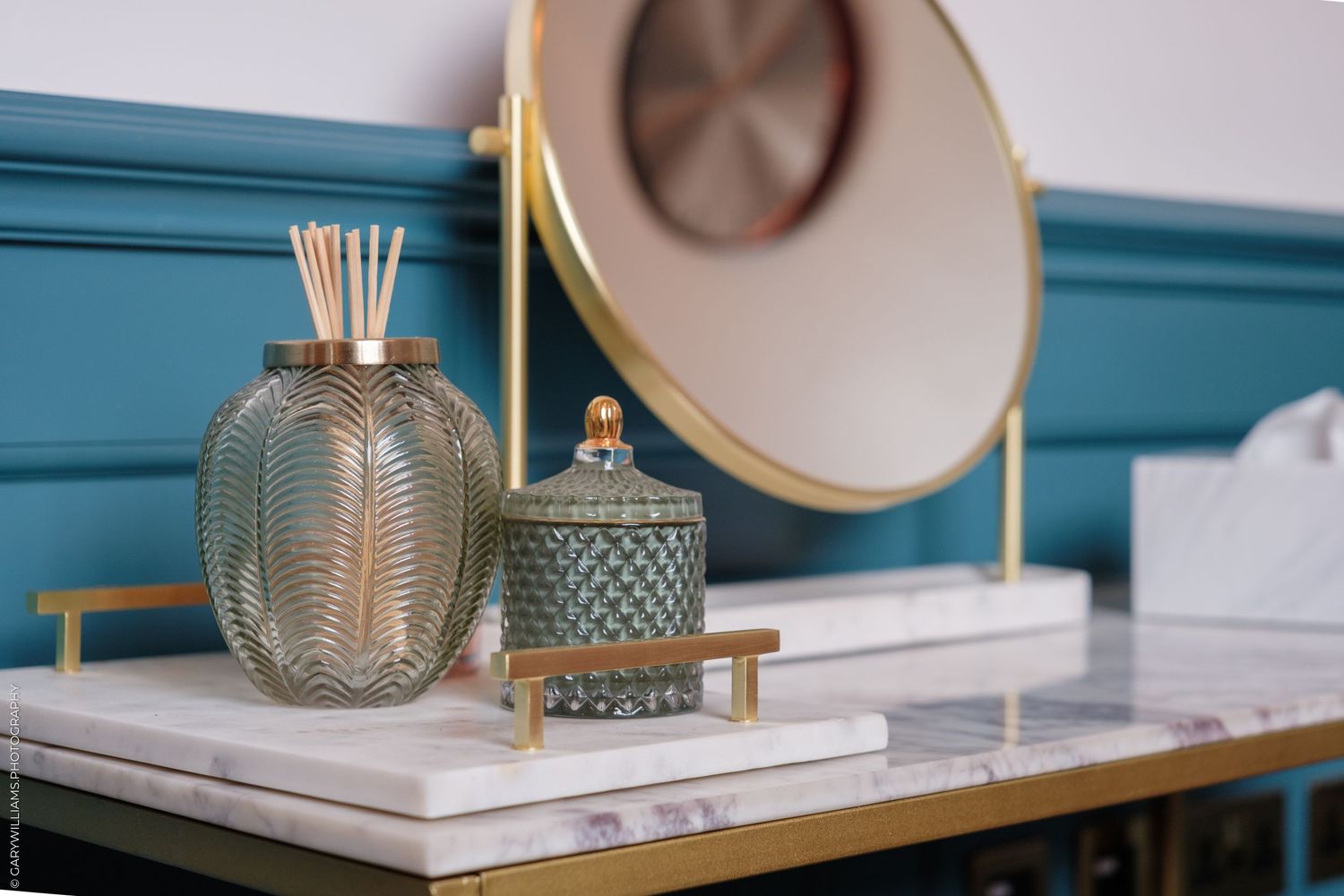 Elegant vanity tray with gold mirror, glass reed diffuser, and decorative jar on marble surface with teal wall.