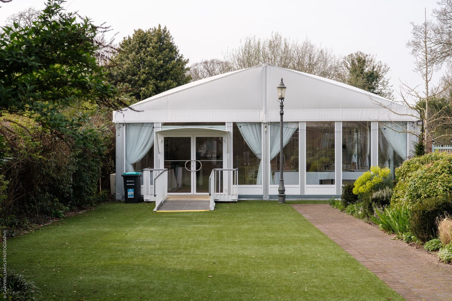 White marquee event venue with glass doors, draped curtains, and manicured lawn in a garden setting.