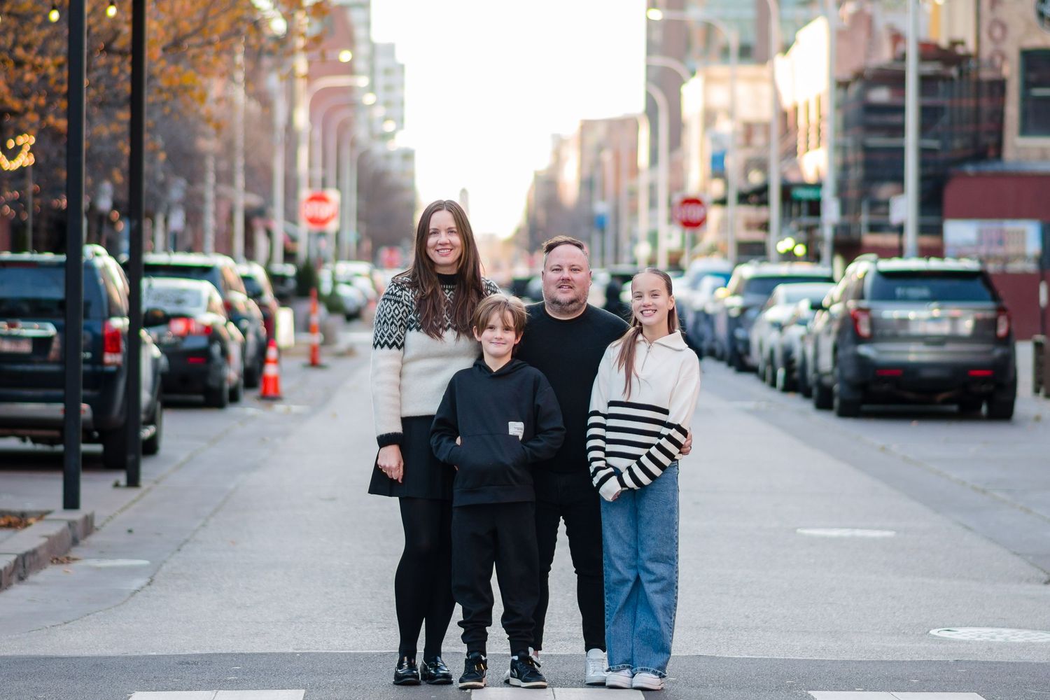 Family of four posing together on a city street lined with parked cars and traffic cones on a fall day.