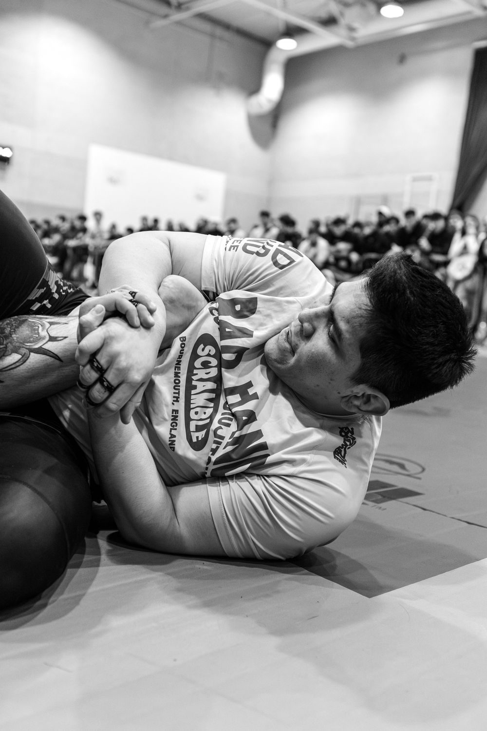 Two BJJ competitors grappling on the mat during a submission wrestling tournament in a crowded gym.