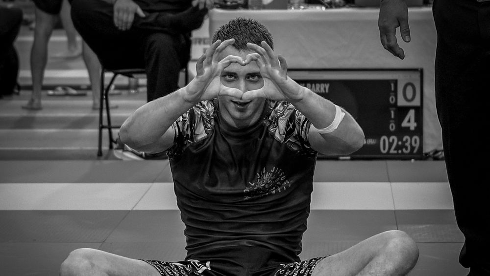 BJJ athlete sits cross-legged on the mat celebrating victory with hands on head at a tournament.