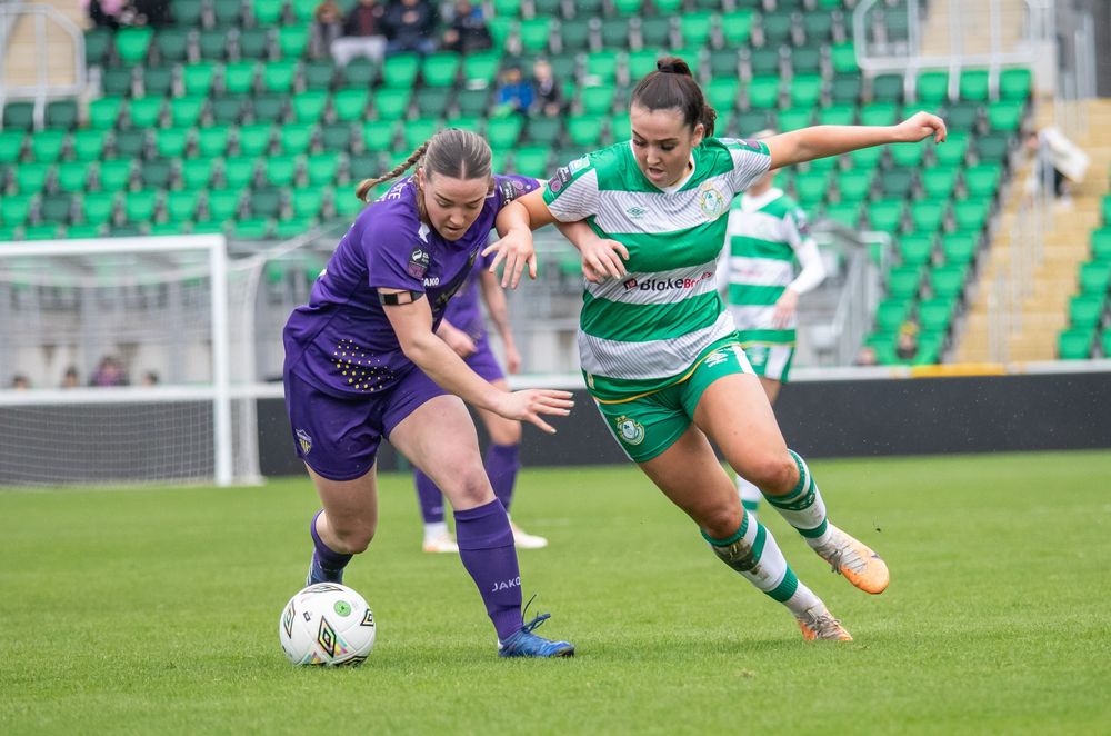 Two women footballers battle for possession during a competitive match at a green stadium.